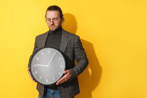 Sad man wearing glasses holding big clock on yellow background