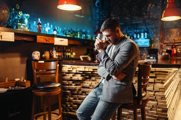 Sad man with glass of alcoholic beverage at wooden bar counter. Tired customer in pub, male person relax in restaurant