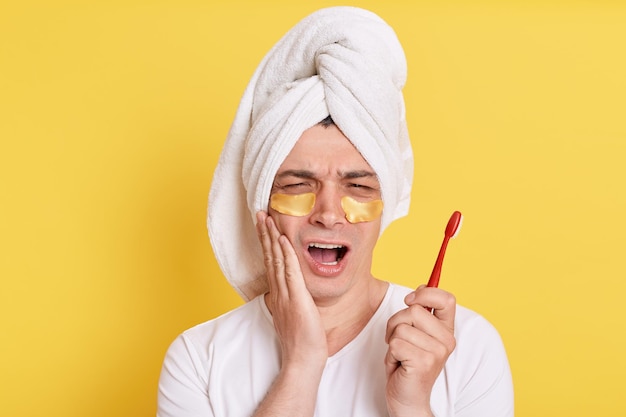 Sad man wrapped towel on head doing morning cosmetology procedures posing with patches under eyes isolated over yellow background holding brush in hands feels pain after brushing