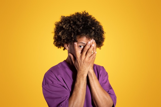 Фото Грустный взрослый африканский кудрявый мужчина в фиолетовой футболке закрывает лицо руками на желтой студии