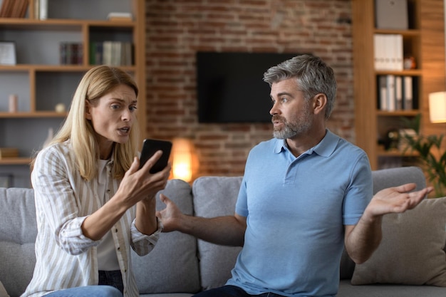 sad mature caucasian wife shows smartphone to shocked husband man suffers from gadget play addiction
