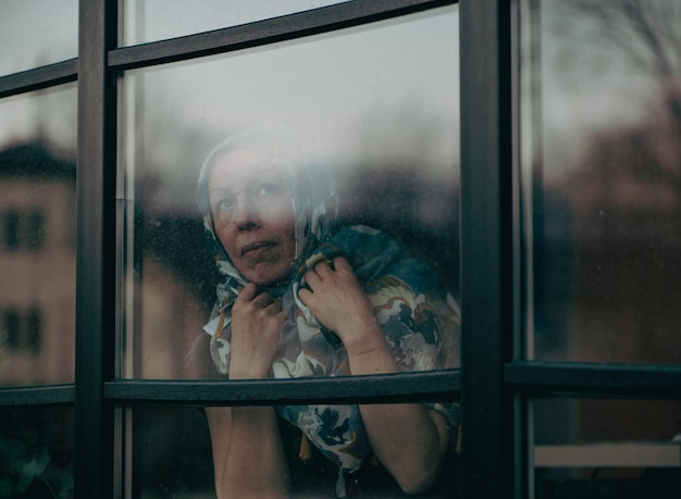 汚れた窓ガラスの後ろに立って頭に scraft を持つ悲しい成熟した女性