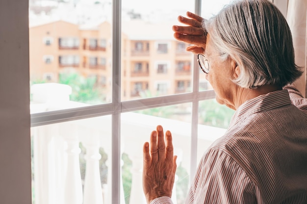 Sad melancholy senior caucasian woman at home looking out the window pensive