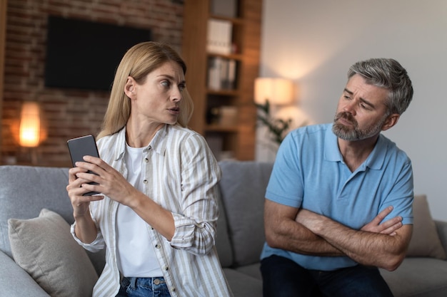 sad middle aged european man looking at scared woman with smartphone in living room interior