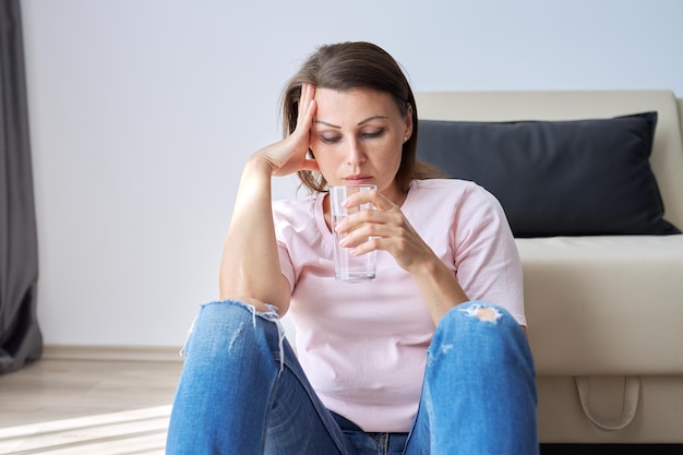 Foto triste donna di mezza età seduta a casa sul pavimento con un bicchiere d'acqua problemi di salute delle donne anziane salute mentale dolore emicrania menopausa sintomo depressione periodo dopo coronavirus covid19