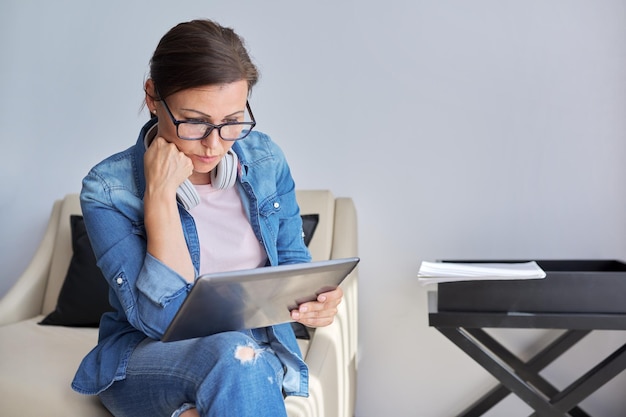 Triste donna di mezza età con tavoletta digitale seduta sulla poltrona a casa