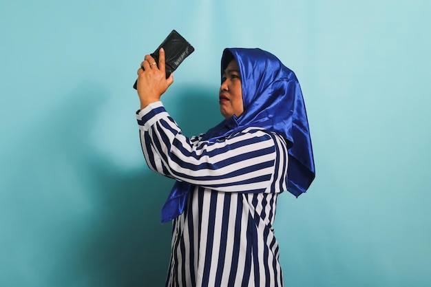 Photo sad middleaged asian woman in hijab is looking into her empty wallet isolated on blue background