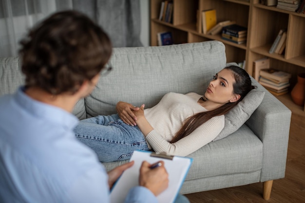 Sad millennial european woman suffering from mental problems lies on couch talk with man doctor in clinic