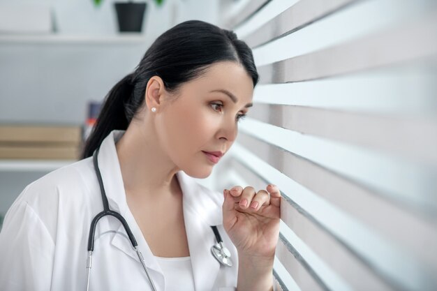 Sad mood. Successful pretty adult woman in a white medical coat, standing near the window, resting her head on the blinds.