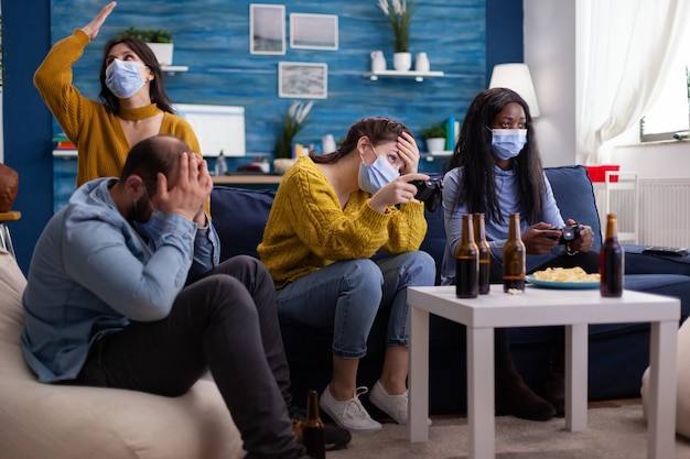 Sad multi ethnic group of friends after lose at gaming competiton with face covers with mask during covid outbreak keepin social distancing in home living room. enjoying beer and chips. conceptual ima