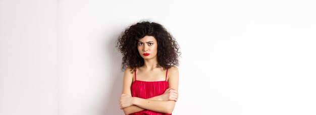 Sad and offended caucasian woman in red dress frowning cross arms on chest and sulking feeling mad a