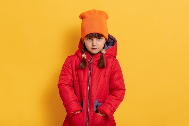 Sad offended little girl wearing orange cap and red jacket standing against yellow wall and looks at front with upset facial expression