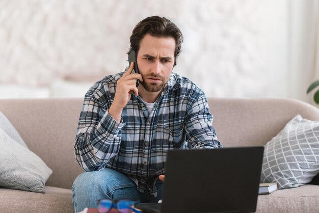 Sad pensive millennial european guy in domestic clothes with stubble calls by phone look at computer