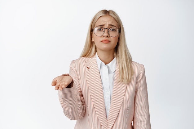 Sad poor corporate woman saleswoman raising empty hand begging for money with miserable face expression standing in suit and glasses over white background