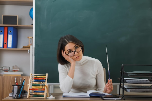 sad putting hand pn cheek young female teacher wearing glasses holding pointer sitting at desk with school tools on in classroom