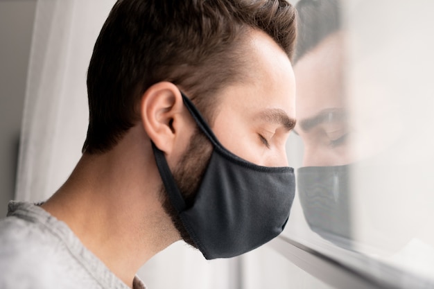 Sad puzzled young man in cloth mask leaning head on window and keeping eyes closed while feeling depression because of coronavirus isolation