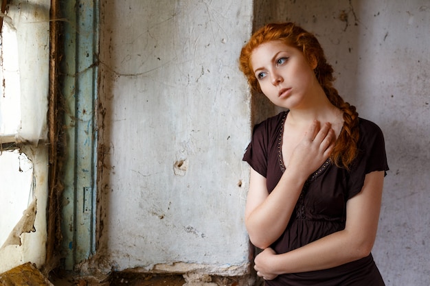 Sad red-haired girl standing near a broken window, the concept of poverty and misery