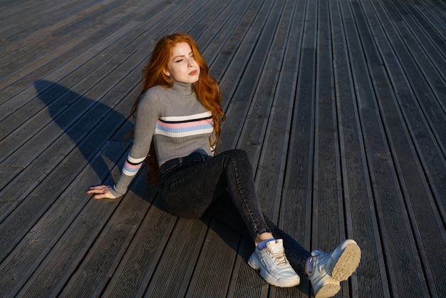 Sad redhead girl sits on a wooden floor in the park on a sunny day