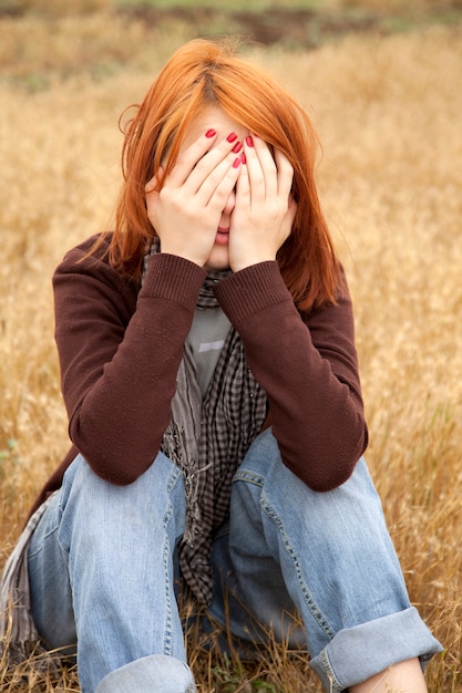 Sad redhead girl sitting on yellow grass at countryside 