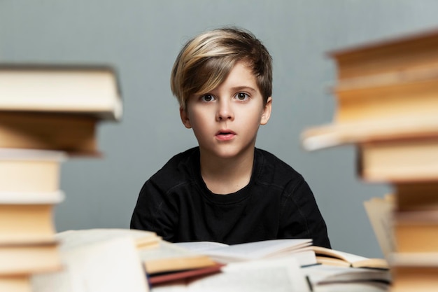 A sad schoolboy sits at a table with a pile of books Cute boy in a black tshirt Difficulties in learning and stress Grey background