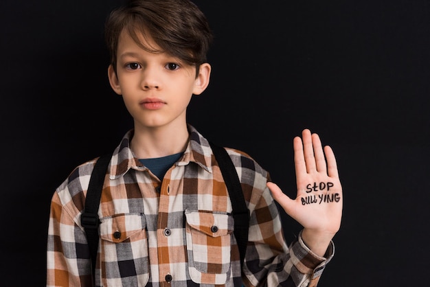 Sad schoolboy with stop bullying lettering on hand isolated on black