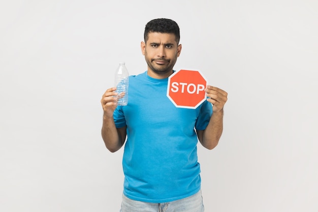 Sad serious unshaven man holding red stop symbol and empty plastic bottle earth protection