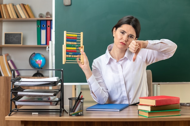 Sad showing thumb down young female teacher sitting at table with school tools holding abacus in classroom