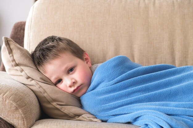 Грустный больной мальчик лежит на диване у себя дома под синим одеялом