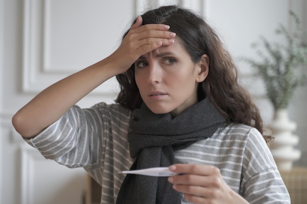 Sad sick young Italian woman wearing knitted scarf around neck suffering from flu cold at home