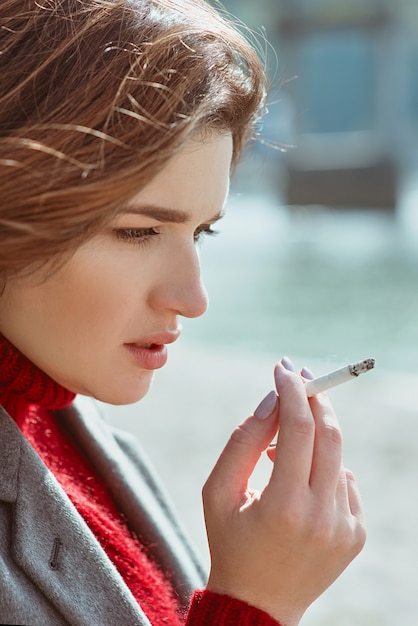 sad stressful young woman in sweater and coat smoking cigarette near the river