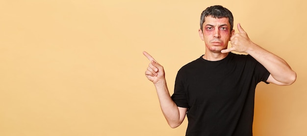 Грустный страдалец в черной футболке с синяками и ссадинами на лице, изолированный на бежевом фоне, указывающий на копировальное пространство, показывающее телефон доверия