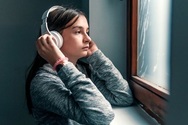 Sad teenage girl sitting on windowsill with earphones listening to music