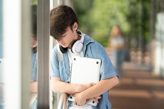 Sad teenager boy holding laptop against wall in a corridor