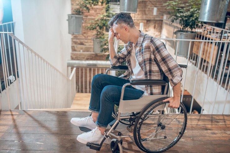 Sad thinking. Adult sad man on wheelchair in thought touching his face with fist standing near stairs down in room