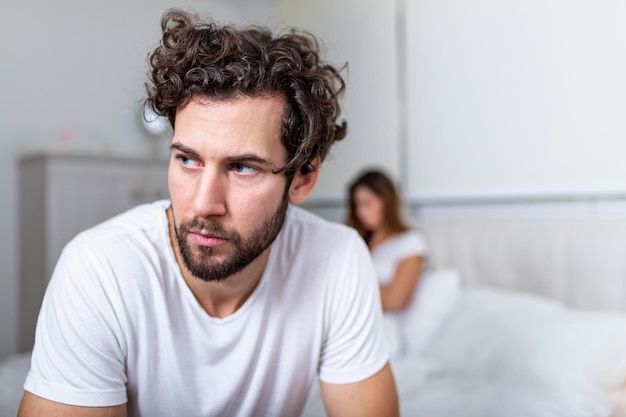 Sad thoughtful man after arguing with girlfriend.Relationship difficulties, conflict and family concept,unhappy couple having problems at bedroom. Sad guy sitting on bed,girlfriend in the background