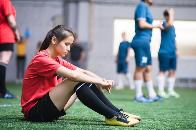 他のチームの緑のフィールドに座っているスポーツユニフォームの悲しいまたは疲れた女性のサッカー選手