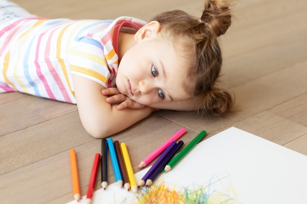 Sad tired frustrated bored stressful child lies on wooden floor\
with white sheet of paper and colored pencils. learning\
difficulties, education concept. family relationship. misses\
home