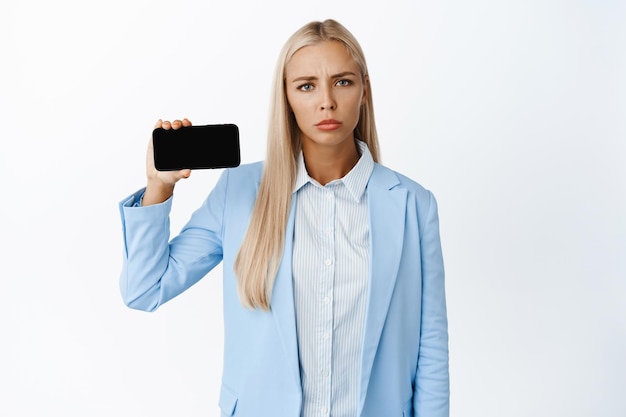 Sad and tired young businesswoman showing smartphone screen horizontally standing in blue suit over white background