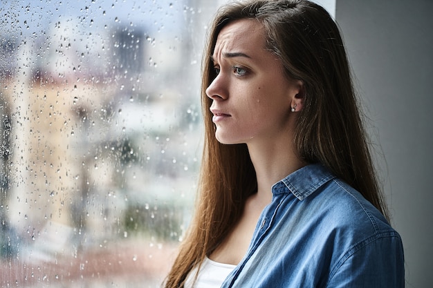 Sad unhappy crying woman with tears eyes suffering from emotional shock, loss, grief, life problems and break up relationship standing by the window with raindrops. Female received bad news