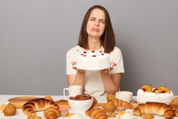 회색 배경 위에 격리된 흰색 티셔츠를 입은 슬픈 화가 난 매력적인 여성은 맛있는 케이크를 들고 있는 집에서 만든 디저트들 사이에서 축제 테이블에 앉아 있습니다.