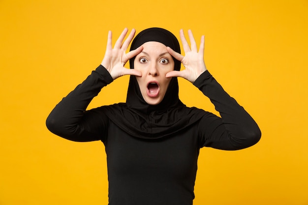 Sad upset crying confused young arabian muslim woman in hijab black clothes posing isolated on yellow wall, portrait. People religious Islam lifestyle concept. 