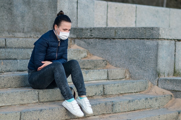 悲しい動揺して落ち込んでいる女の子、階段の上に座っている孤独な欲求不満な若い女性、隔離のために苦しんでいる、コロナウイルス。顔に医療用防護マスクの人。失恋、ウイルス、流行のコンセプト