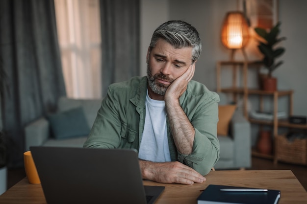 Sad upset elderly european male with beard watches video on computer at table in room interior