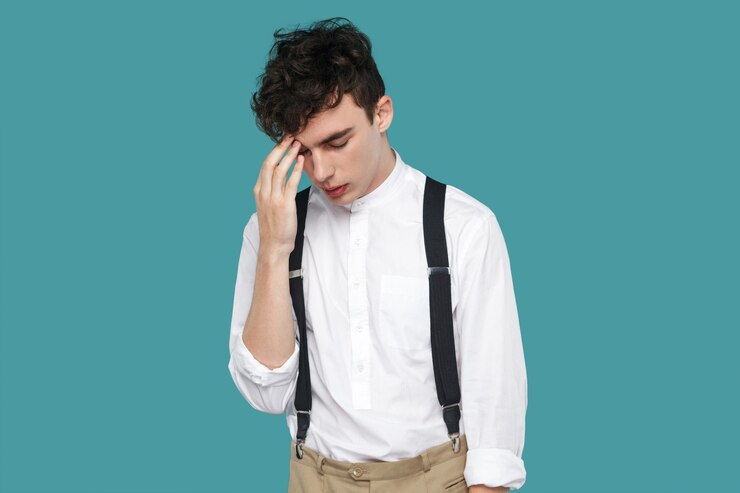 Photo sad upset man holding head down and thinking. portrait of handsome hipster curly young businessman in classic casual white shirt and suspender standing. indoor studio shot isolated on blue background.