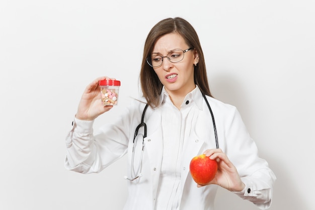 Sad upset pretty young doctor woman with stethoscope, glasses isolated on white background. Female doctor in medical gown holds bottle with pills, red apple. Healthcare personnel, medicine concept.