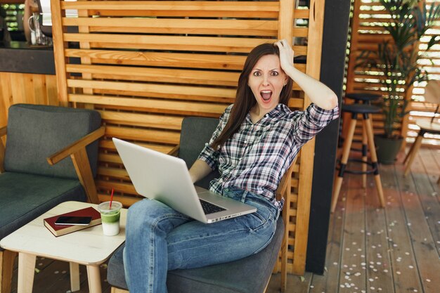 Photo sad upset screaming woman in outdoors street summer coffee shop sitting in casual clothes, working on modern laptop pc computer during free time