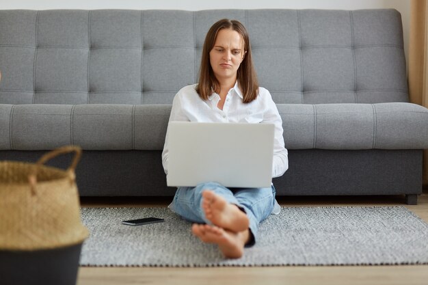 Sad upset tired female wearing white shirt and jeans sitting near sofa on the floor, looking at portable computer screen and expressing sadness, freelancer does not want to work.