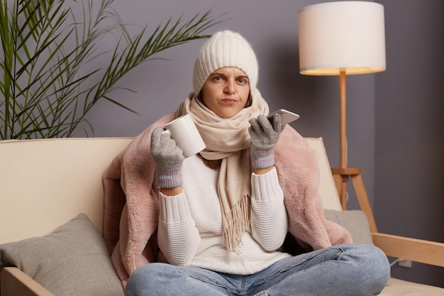 Photo sad upset woman wearing warm clothes and wrapped in scarf sitting on sofa in room with cup of hot tea in hands and cell phone trying to get through to the master to fix the heating in the house