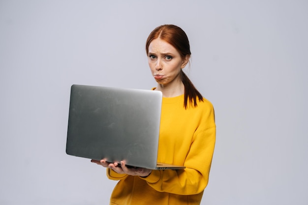 Грустная расстроенная молодая деловая женщина или студентка, держащая открытый ноутбук и смотрящая в камеру на изолированном сером фоне