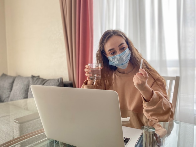 Sad, upset, young woman holding a glass of water and pills sitting in the living room, feeling ill